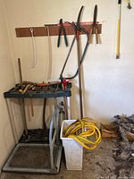 Overview of gardening trolley with tools laid on top, garden hose in container nearby, and hand tools hanging on wooden wall rack.