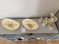 Two oval Wedgwood serving bowls on shelf with several white ceramic pitchers and a sugar bowl arranged beside them