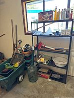 View of entire area showing green plastic wheelbarrow filled with items, black plastic shelving unit holding bottles and gardening supplies, long-handled gardening tools leaning against wall, and other miscellaneous gardening items.