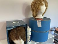 Two wigs displayed with their matching blue storage cases; short brown wig in upright rectangular case and blonde wig on mannequin head atop a larger blue oval case; includes bottle labeled wig cleaner.
