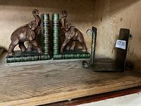 Bronze elephant-themed bookends on left side with stacked book design green base and plain metal bookend on right side on wooden shelf