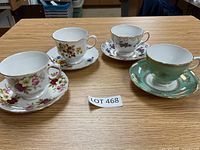 Four bone china teacups and matching saucers displayed on a wooden table, each with distinct floral patterns and gold trim.