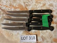 Five serrated steak knives with black plastic handles lying on fabric, grouped and held with green tape on one handle.