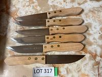 Six steak knives with wooden handles and serrated blades laid out on a marble surface, showing full knife length and knife details.