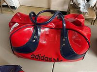 Photo of red Adidas athletic bag on table showing full exterior with navy blue straps and black Adidas logo