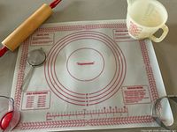 Large Tupperware pastry mat with red printed measurement rings and baking info, accompanying rolling pin, metal spoon, and measuring cup placed on a grey surface.