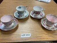 Four bone china tea cups and saucers placed on a wooden table, showing a variety of floral and pastel designs with gold trim, tagged with LOT 455.