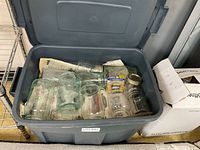 Top-down view of multiple clear glass canning jars in a gray plastic bin with some metal lids and boxes for lids, on top of newspapers.