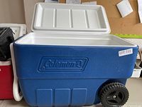 Front view of Coleman cooler showing blue body, white lid, brand logo, handle on one side, and one black wheel visible.