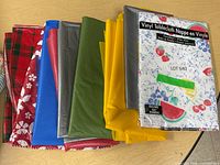 Nine plastic and vinyl tablecloths folded and laid out, showing various solid colors and patterns.