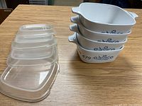 Photo of four square Corning Ware baking dishes stacked with blue cornflower design and four clear glass lids beside them on a wooden surface.