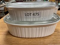 Two stacked white ribbed ceramic Corning Ware casseroles with clear lids, labelled Lot 475 and Lot 476 visible.