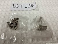 Pair of Transformers Autobot logo cufflinks in silver with black detail, placed on a white textured cloth.