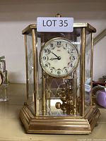 Front view of Kundo mantel clock showing floral clock face, brass details, and rotating pendulum balls inside glass case.