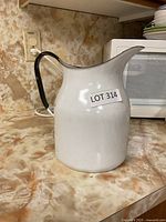 Side view of white enamel ware pitcher with black handle and spout on a marble countertop.