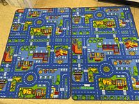 Two rectangular blue floor mats with colorful road and city designs, placed side by side on a yellow tiled floor.