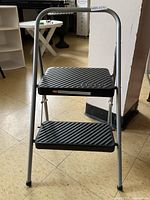 Full front view of the two-step folding stool showing metal frame and black plastic steps with textured diagonal grip patterns.