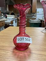 Front view of pink glass vase on wooden table with kitchen background. Vase is 6 inches tall with spiral clear glass decoration on neck and flared, ruffled rim.