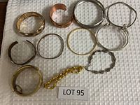 Top down photo of 11 bracelets in gold and silver tone on white textured cloth, showing various styles and designs of bangles, cuffs, and chain link bracelets neatly arranged.