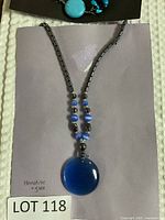 Hematite and glass bead necklace with large round blue pendant placed on a beige surface with handwritten note.