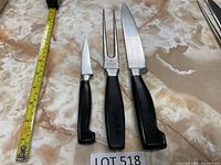 Three Henckels cutting tools: paring knife, carving fork, and chef's knife, arranged side by side on beige marble surface with measuring tape nearby for scale.