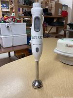 Full view of KitchenAid immersion blender standing upright on a wooden table with background shelves and other items.