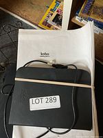 Kobo eReader in black case with USB cable on Kobo-branded white cloth background. Elastic band securing the case closed with lot sticker on front.