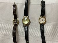Three ladies' watches laying flat on fabric with visible brown and black leather straps, round faces, and varying dial colors including dark, gold-tone, and white.