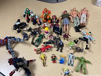 Overview of assorted action figures and toys spread on a wooden surface, includes figures in green, orange, black, and red colors plus smaller figures and novelty pieces.