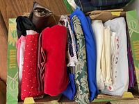 Box filled with folded assorted fabrics showing different colors and textures such as red, black, blue, brown, and white, some quilted and some with prints.
