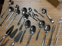 Top view of the majority of serving utensils spread out on a wooden surface. Includes large spoons, spoons with ornate handles, ladles, pie servers, and forks.