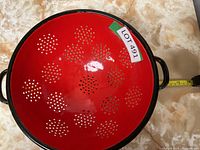 Top-down image showing red metal colander with flower-shaped perforation patterns, black rim, and black side handles.