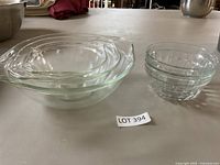 Set of three large clear glass mixing bowls and five smaller ribbed glass bowls shown on a table under light background.