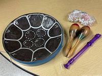 Photo of blue steel tongue drum with note markings, two wooden maracas, purple plastic recorder, and two bags of marbles on a wooden surface.