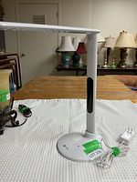 Full view of white OttLite desk lamp showing adjustable flat arm and base with power cord and adapter plugged in.