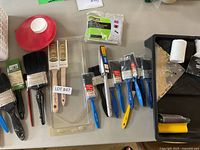 Assorted paint brushes in various sizes, with some in packaging and some unpackaged, paint roller cover, plastic paint tray liner and roller cleaning tool