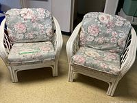 Two white wicker armchairs each with floral upholstered cushions, photographed together in a basement setting.