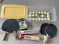 Five ping pong paddles and multiple white table tennis balls including boxed new balls shown on surface