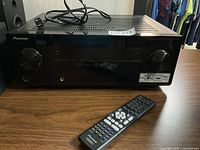 Front view of black Pioneer VSX-821 receiver on wooden surface with power cord on top and remote control in front on table.