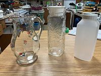 Three pitchers arranged on a wooden table, including two glass pitchers and one plastic pitcher, in a basement setting with kitchen items in background.