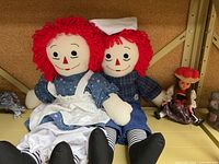Pair of Raggedy Ann and Andy dolls seated side by side with a smaller doll next to them on a shelf