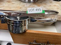 Stainless steel food ricer placed on a counter with LOT 495 tag on handle, showing perforated basket and lever mechanism.