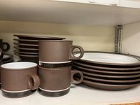 Stacked brown and white bowls and plates along with brown coffee cups on a shelf.