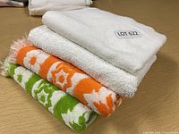 Stack of four towels: two solid white towels on top, one orange floral patterned towel, and one green floral patterned towel beneath, all neatly folded.