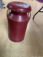 Front-angle image of a red painted metal vintage milk can with lid and handles on beige flooring.
