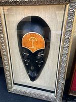 Front view of African carved wooden mask with painted orange forehead mounted in ornate carved frame on light background.