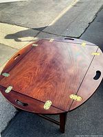Wooden butler's tray table with reddish-brown finish and brass hinges on the top, showing the tray closed with hand grip cutouts.