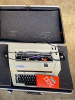 Top-down view of IBM electric typewriter inside open metal carrying case with power cable and ribbon cartridge laid across keyboard