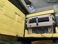 IBM beige electric typewriter placed inside a yellow foam lined carrying case with coiled power cord, viewed from top showing keyboard and paper feed mechanism.