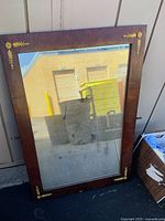 Full view of the medium rectangular mirror with brown wooden frame and gold decorative corner embellishments, showing reflective glass with visible dirt and wear.